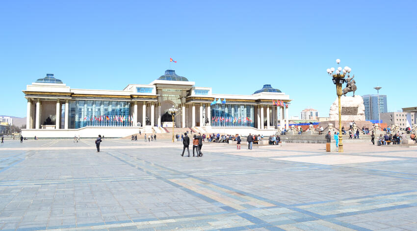 Mongolian Parliament House and Sukhbaatar Square with the Chinggis Khaan monument and people walking on a sunny day.