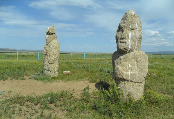 Two ancient stone men (khun chuluu) standing in a fenced grassy steppe under a blue sky at the Ungut complex, Khustai National Park