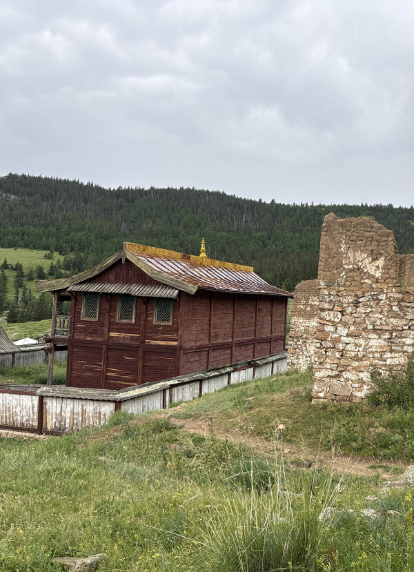 Restored wooden temple beside a stone ruin wall, with forested hills behind, at Manzushir Monastery in Bogd Khan Mountain National Park
